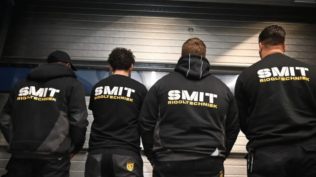 Vier monteurs op de rug gezien, met SMIT Riooltechniek in grote letters op hun zwarte hoodies