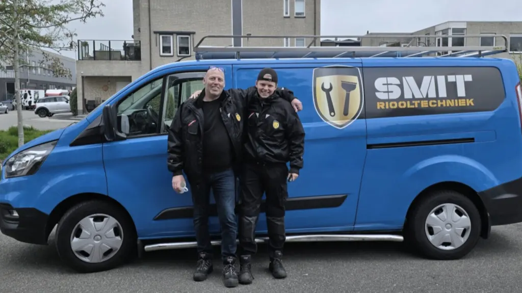 Twee mannen in zwarte SMIT-werkkleding, arm in arm voor een blauwe bus van SMIT Riooltechniek