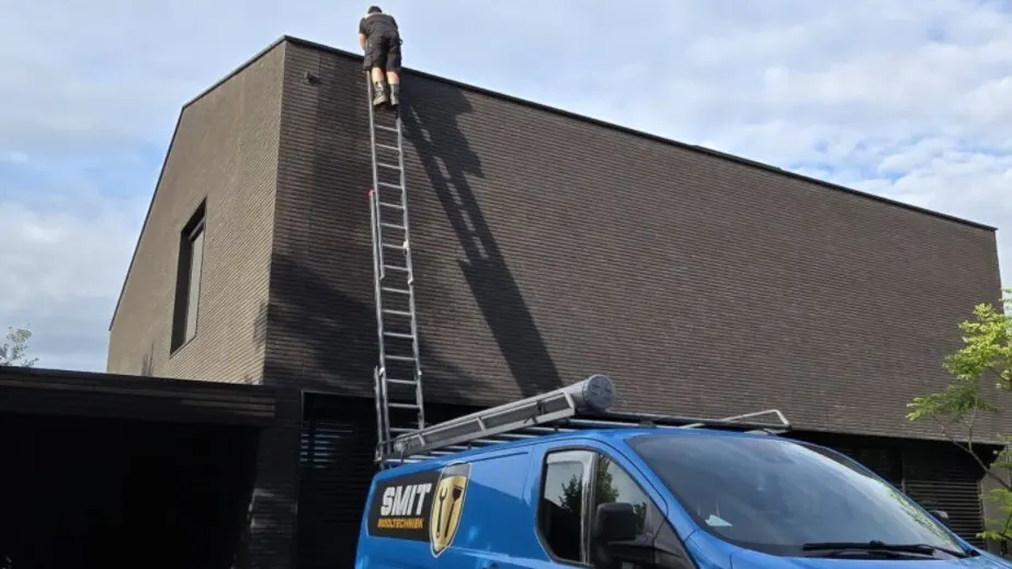 Monteur op een ladder aan de dakrand van een donker gebouw; op de voorgrond een blauwe SMIT-bus