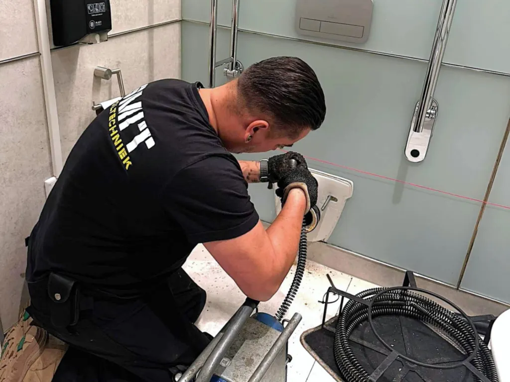 Monteur voert knielend een ontstoppingsveer in een afvoer in een betegelde sanitaire ruimte