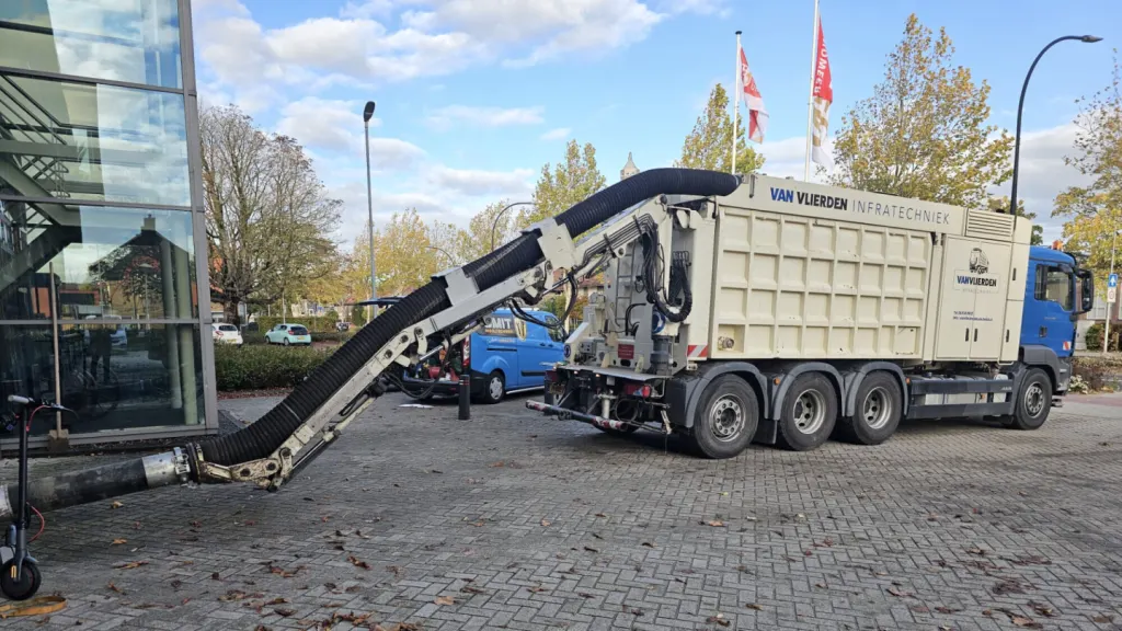 Grondzuigwagen met een lange zuigarm op een bestraat voorterrein; daarachter een blauwe bus