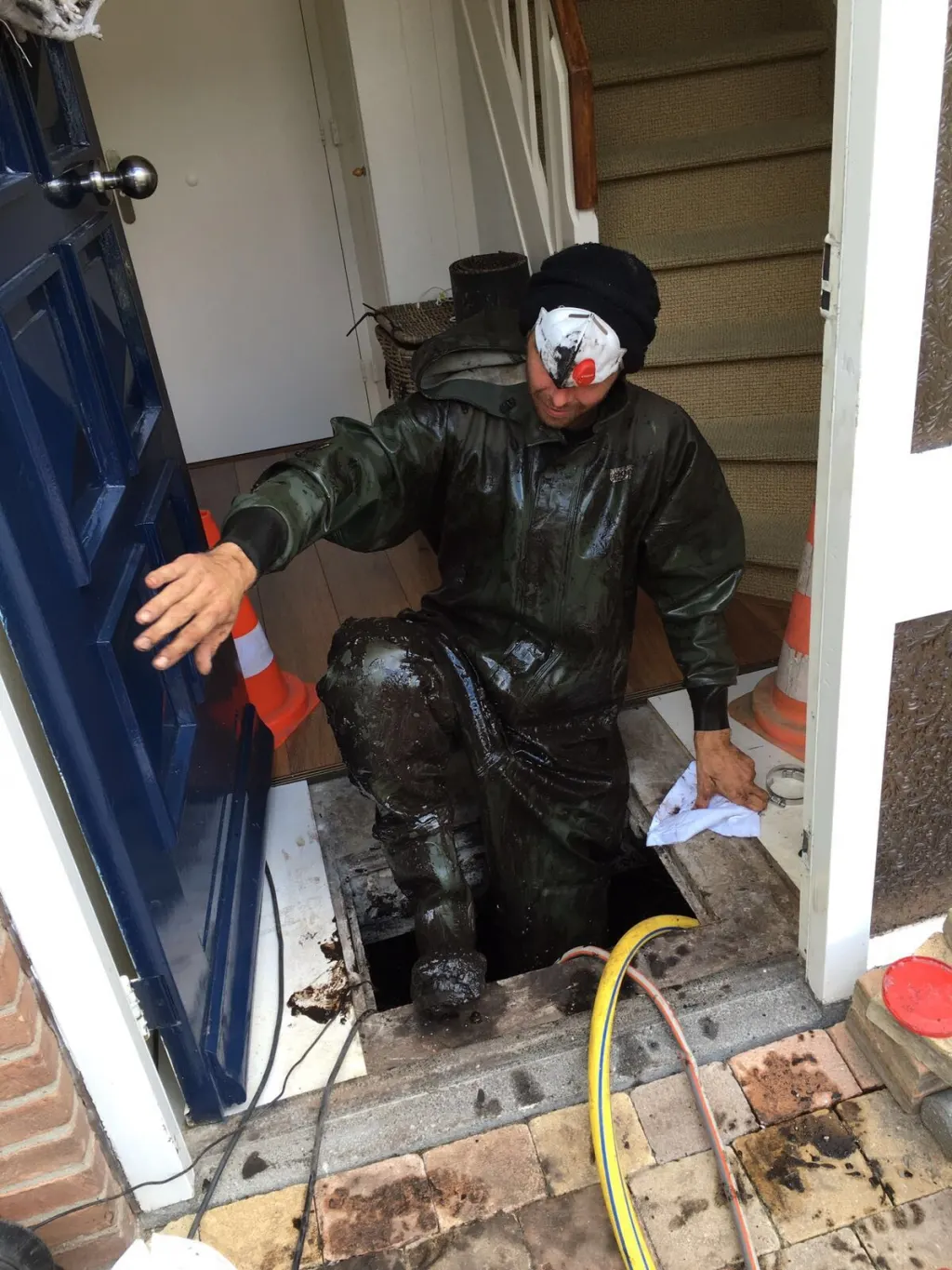 Monteur in een modderig waterdicht pak klimt in een woninghal uit het kruipluik bij de voordeur